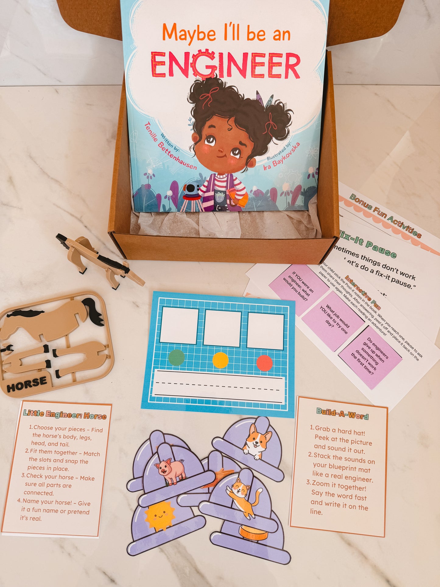 Educational materials including a book titled 'Maybe I'll be an Engineer', wooden horse, and paper cutouts on a marble surface.