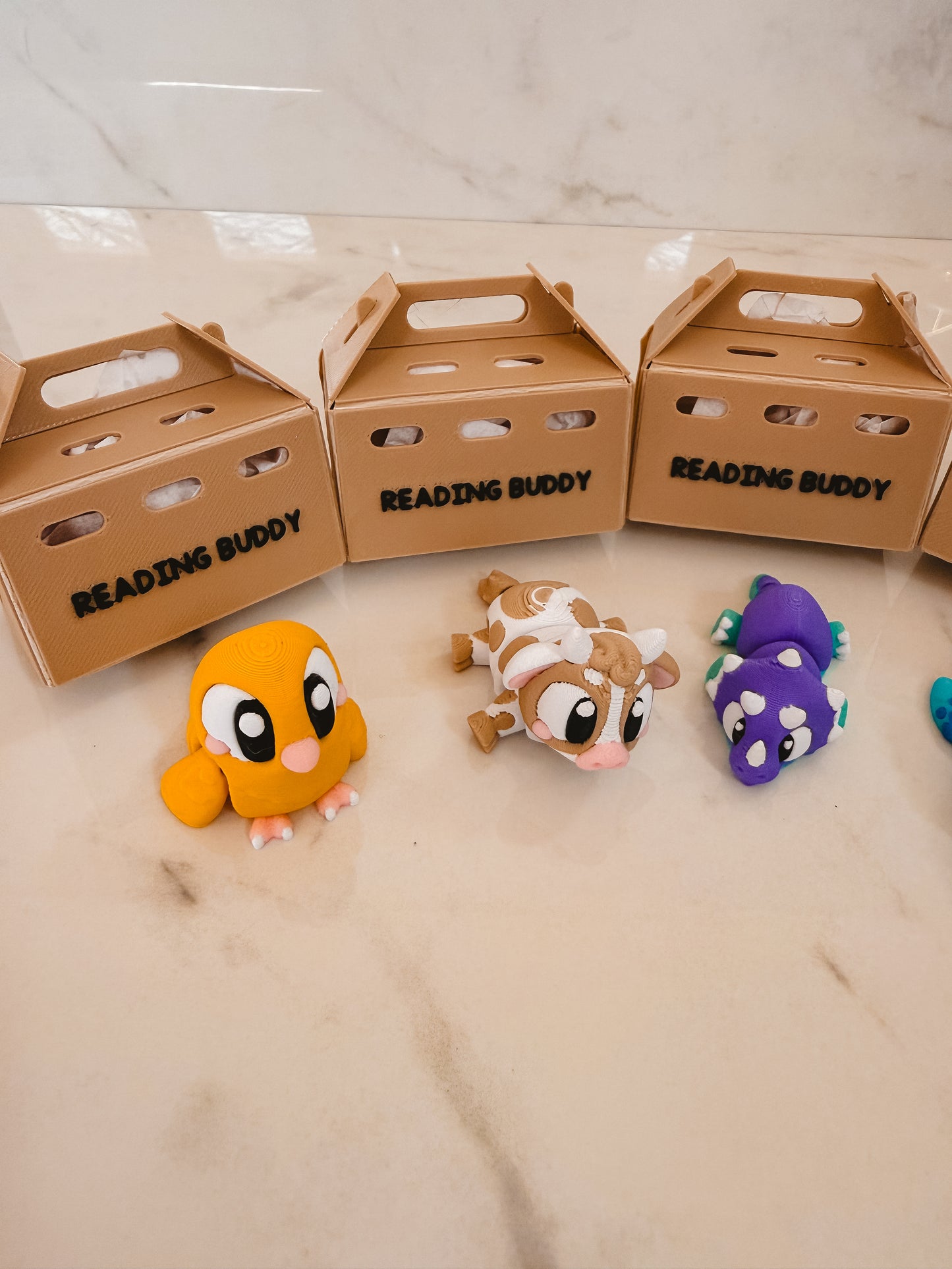 Three cardboard boxes labeled 'Reading Buddy' with colorful animal plush toys on a light surface.