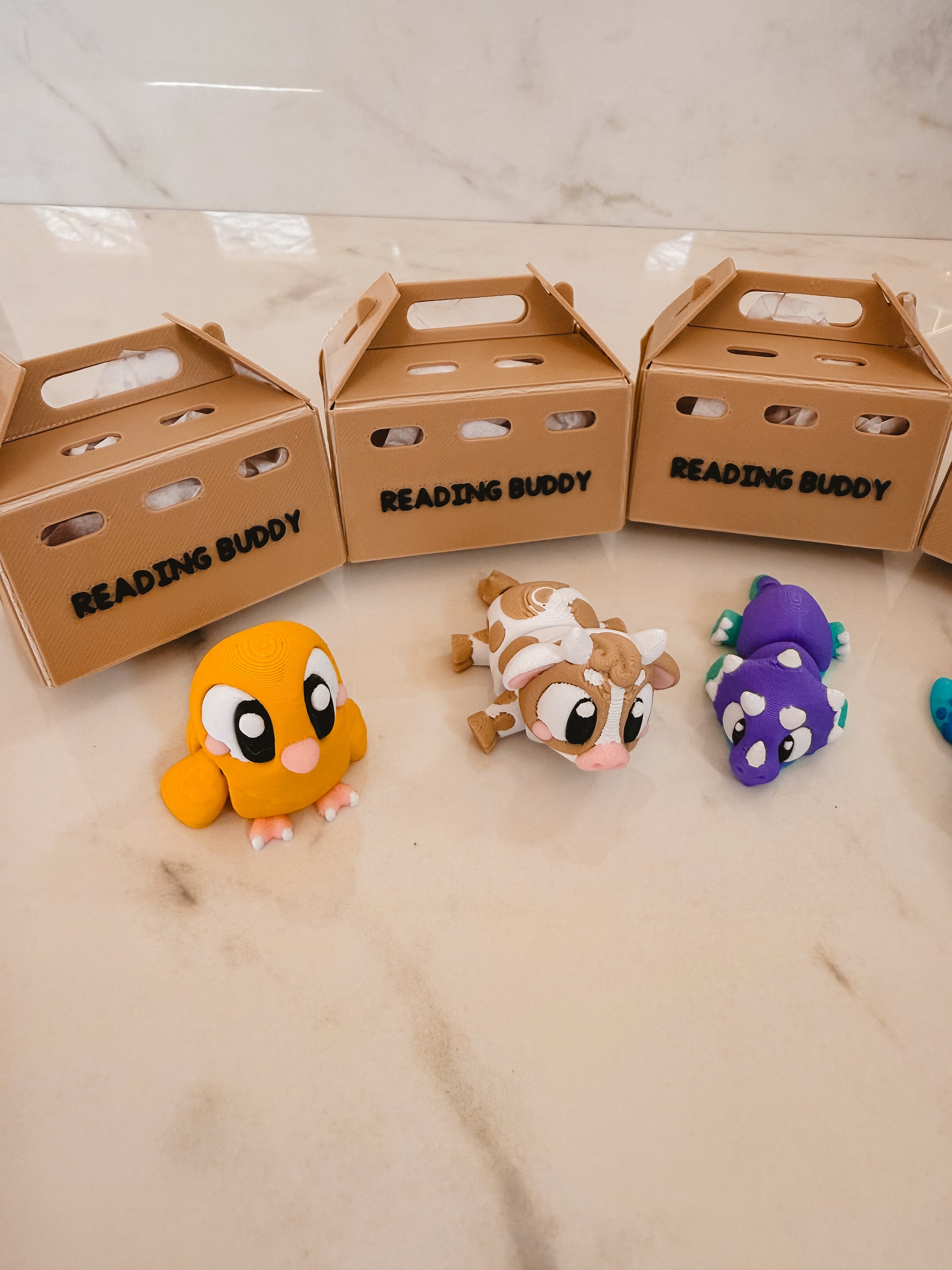Three cardboard boxes labeled 'Reading Buddy' with colorful animal plush toys on a light surface.