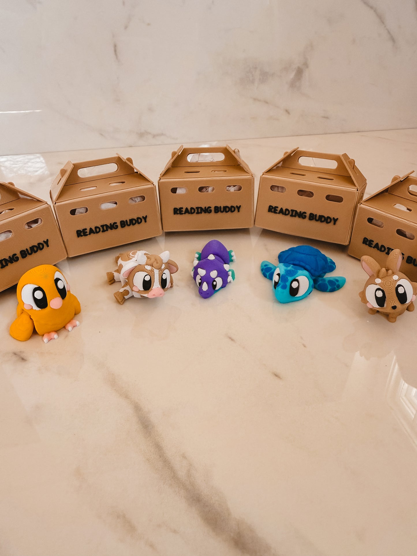 Five colorful plush toys in front of 'Reading Buddy' boxes on a marble surface.