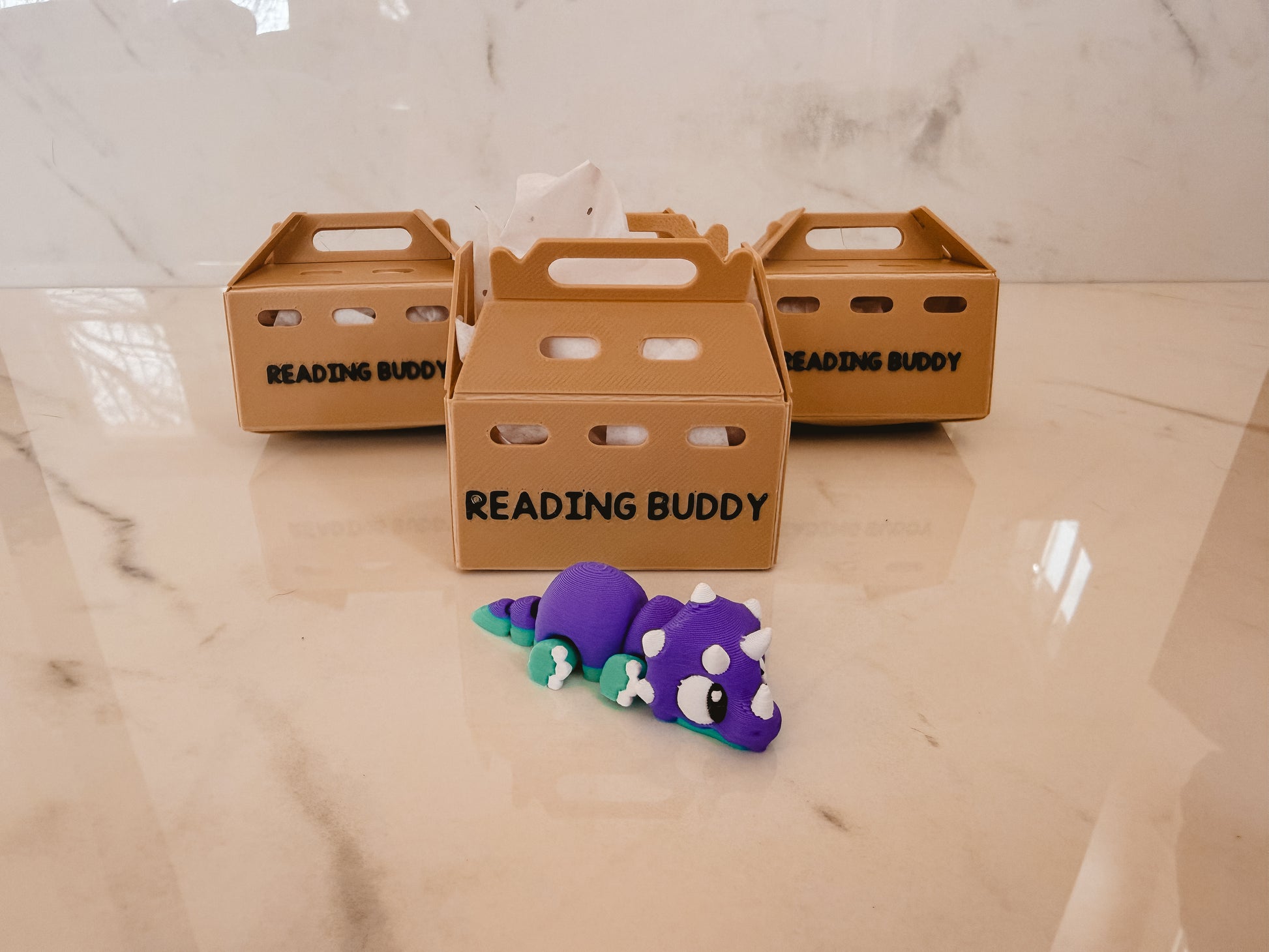 Three cardboard boxes labeled 'Reading Buddy' on a marble surface with a purple toy dinosaur in front.