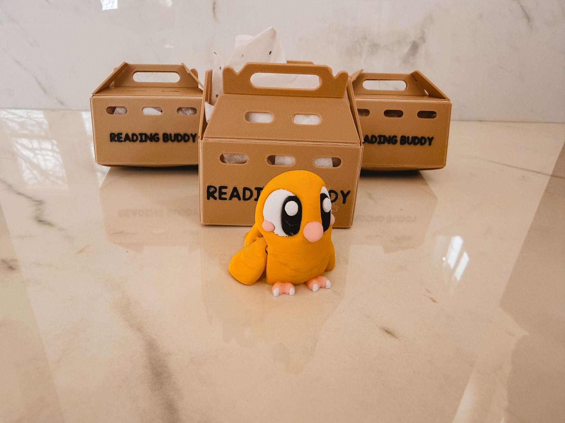 Plush toy resembling a cartoon character in front of 'Reading Buddy' crates on a marble floor.