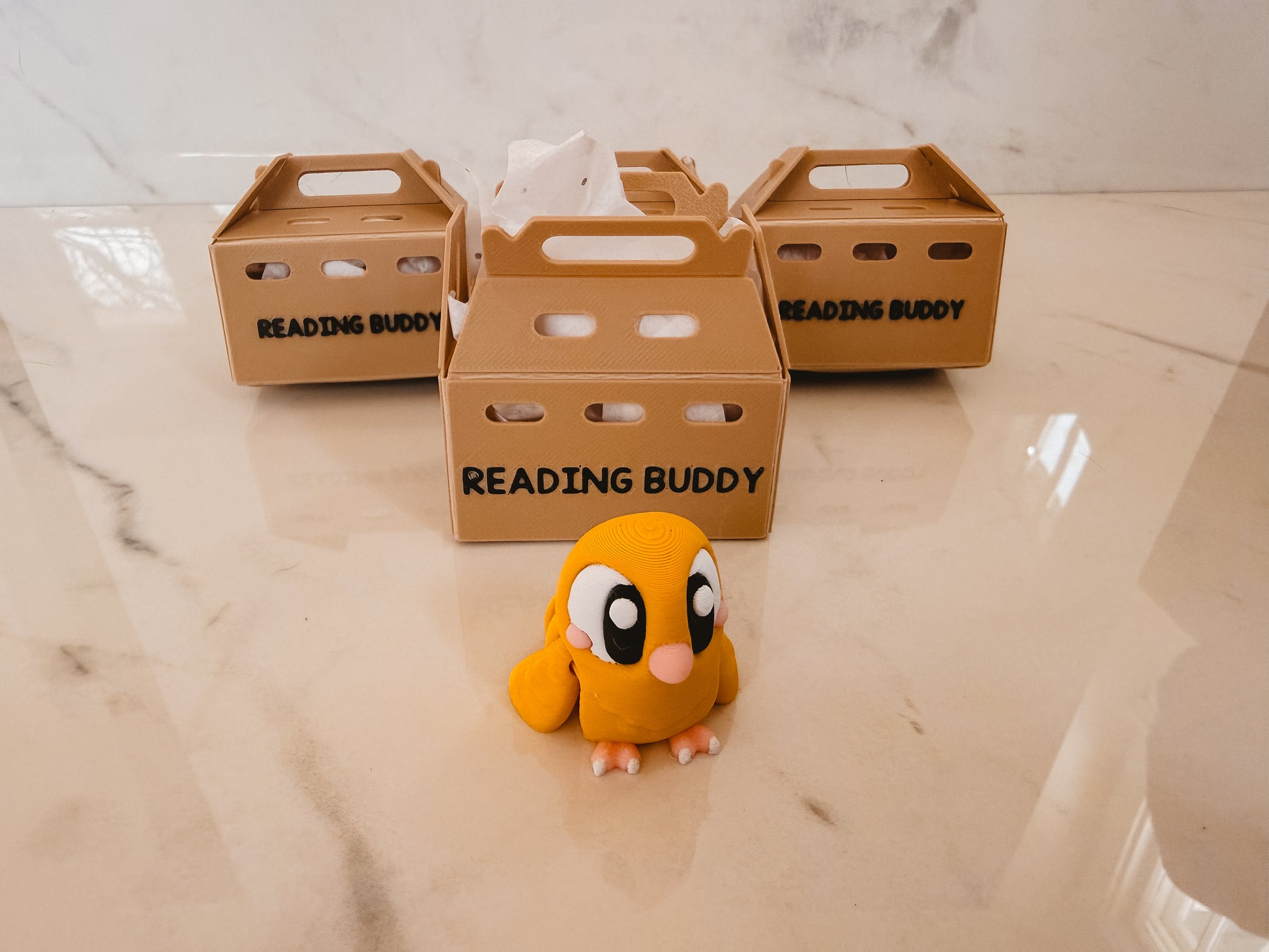 Small orange toy with googly eyes and a pink nose, sitting in front of three cardboard boxes labeled 'Reading Buddy' on a marble surface.