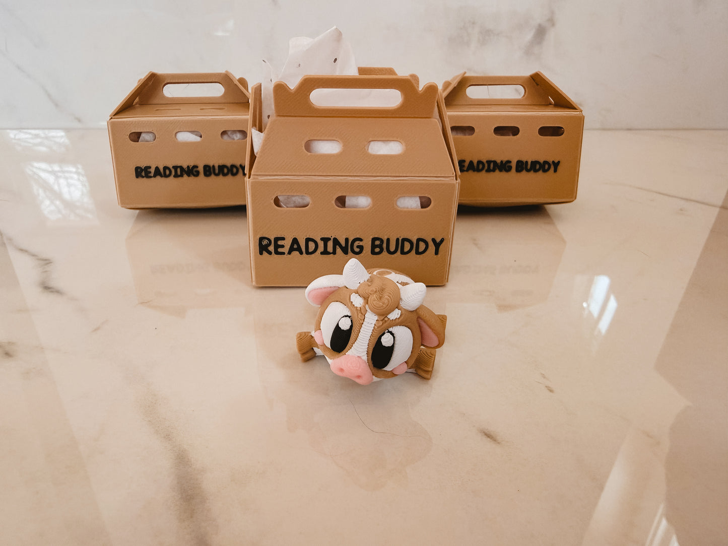 Three cardboard boxes labeled 'Reading Buddy' on a marble surface with a small toy cow in front.