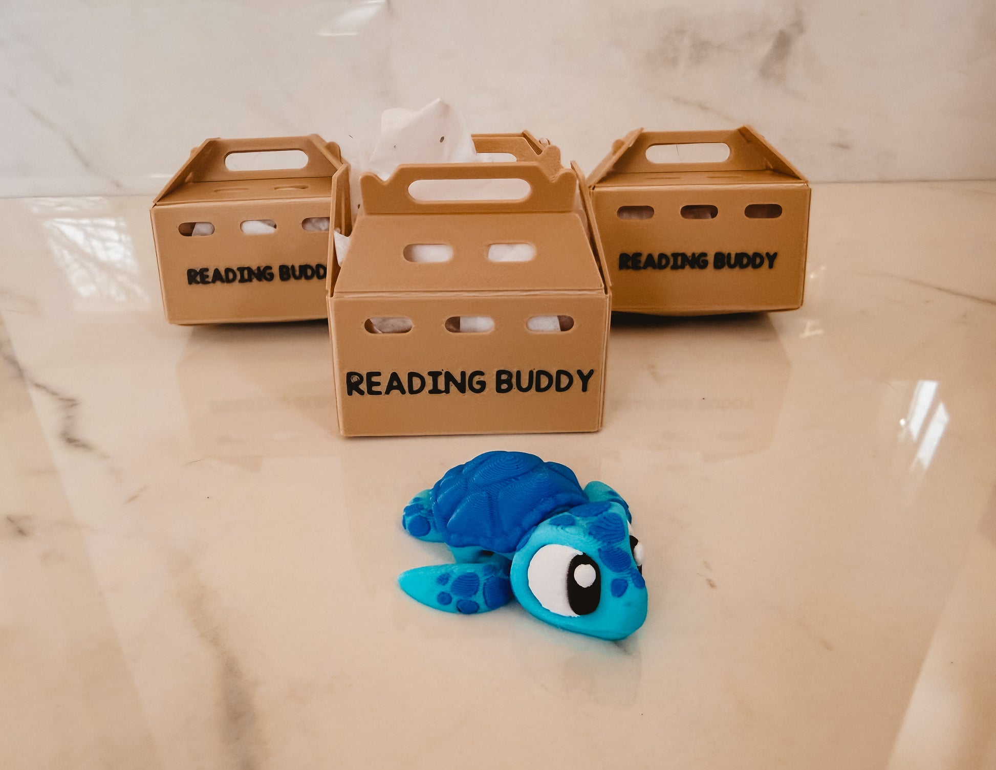 Blue turtle toy labeled 'Reading Buddy' in front of three cardboard boxes with the same label on a marble surface.