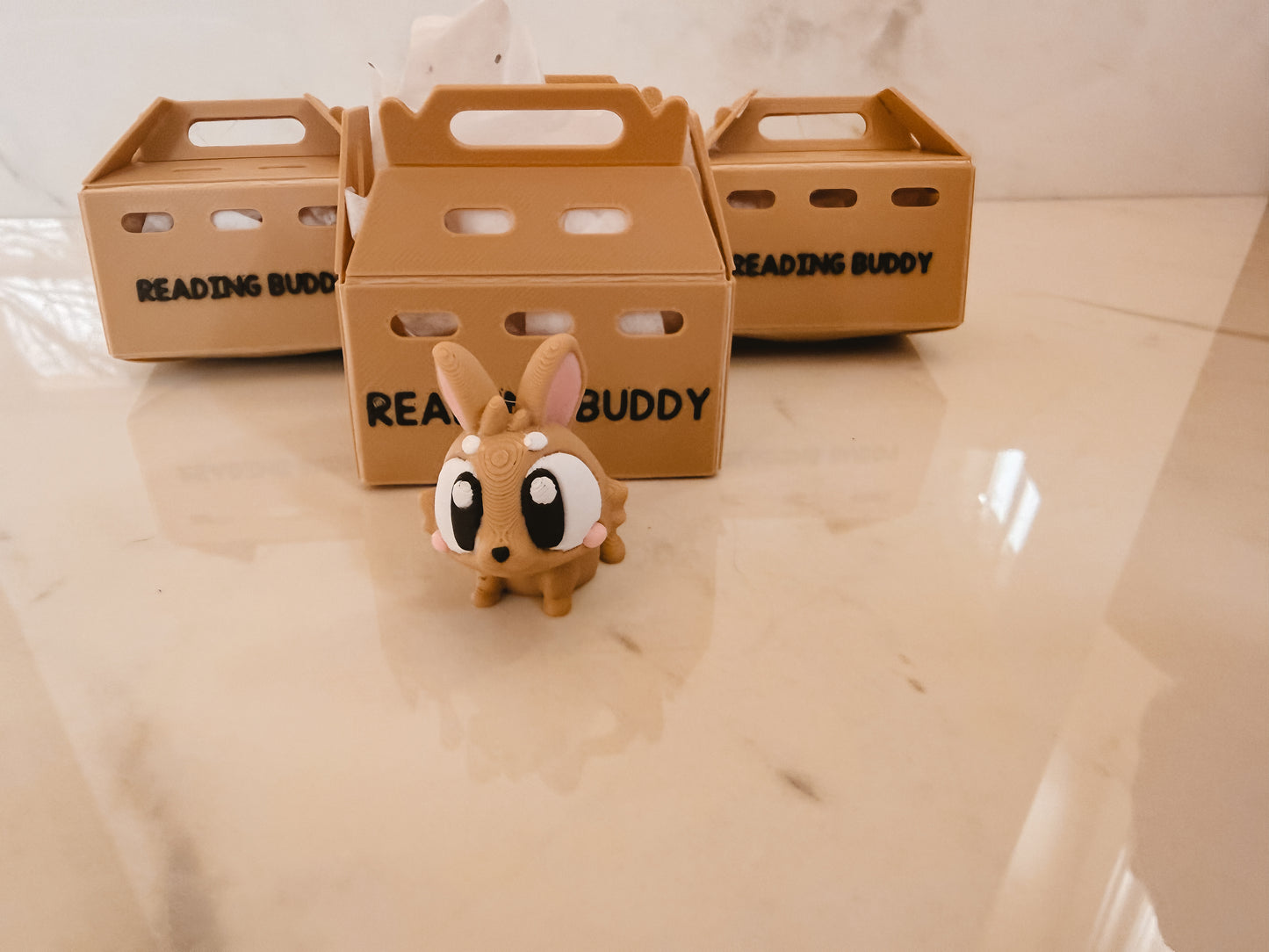 Small brown toy rabbit with googly eyes in front of three cardboard crates labeled 'Reading Buddy' on a light-colored surface.